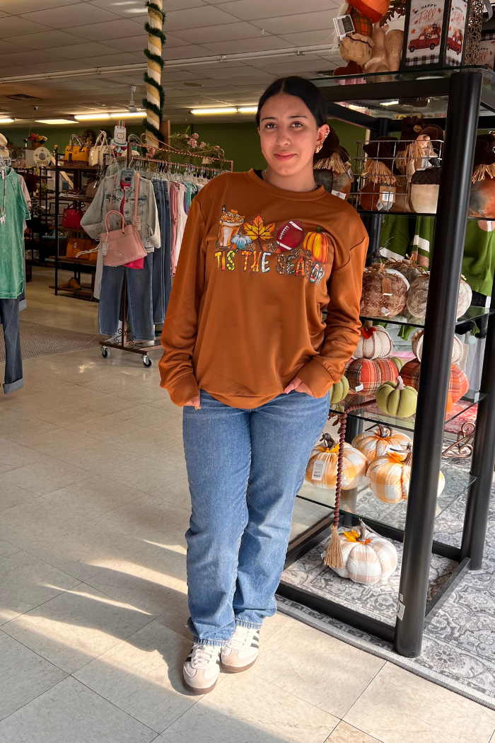 A young woman stands smiling in a boutique, wearing a brown "Tis the Season" sweatshirt with fall-themed graphics like pumpkins and coffee cups, blue jeans, and white sneakers, with clothing racks and decorative pumpkins visible in the background.