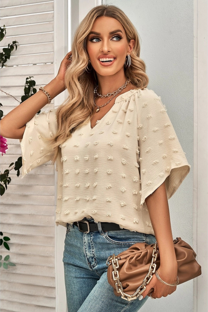 Woman wearing a Swiss Dot Notched Neck Flare Sleeve Blouse. This blouse is a light beige color with small, raised dots all over. It has a V-neck and short, flared sleeves. The woman is also wearing jeans, a black belt, and a gold necklace. She is carrying a brown purse.
