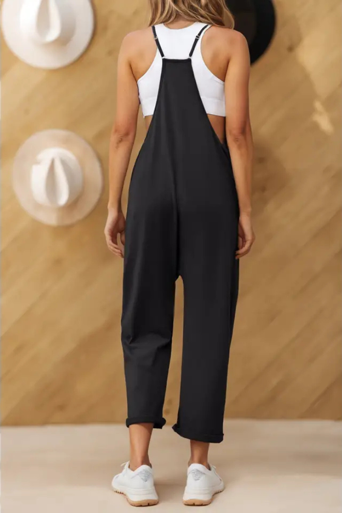 woman from behind wearing a black, sleeveless jumpsuit with spaghetti straps and pockets. The jumpsuit appears to be made from a loose, comfortable material, and the woman is also wearing a white top and white sneakers. In the background, there is a wooden wall with several hats hanging on it. This type of jumpsuit is often described as a casual summer outfit, with features such as a loose fit, pockets, and adjustable straps.