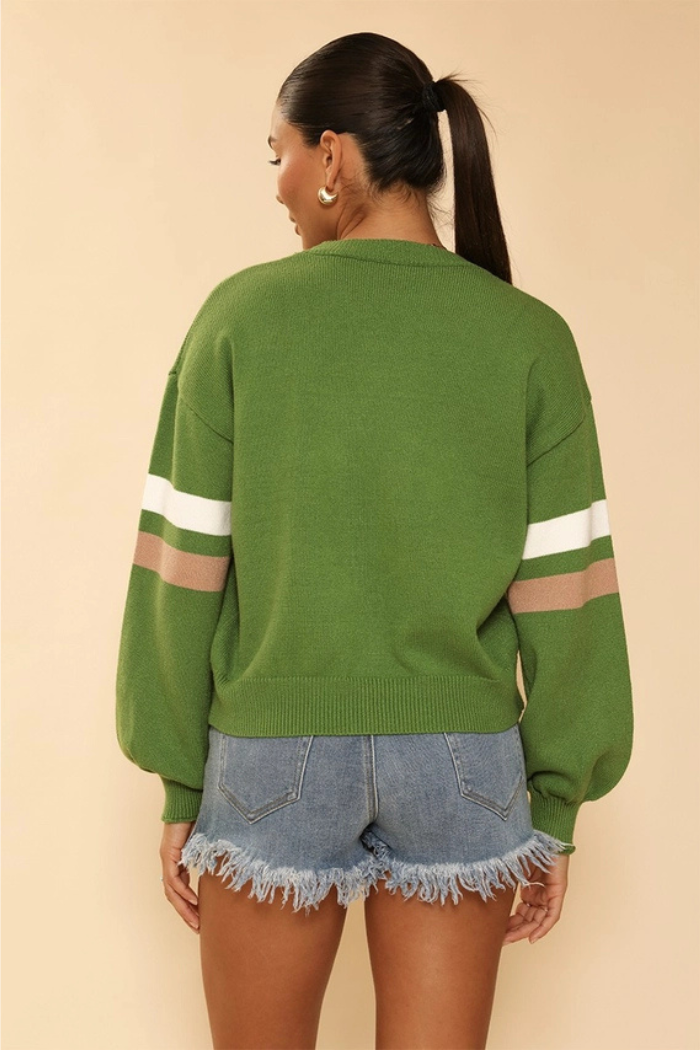 A woman from the back wearing a green sweater with white and brown stripes on the sleeves, paired with distressed denim shorts. Her hair is pulled back in a ponytail.