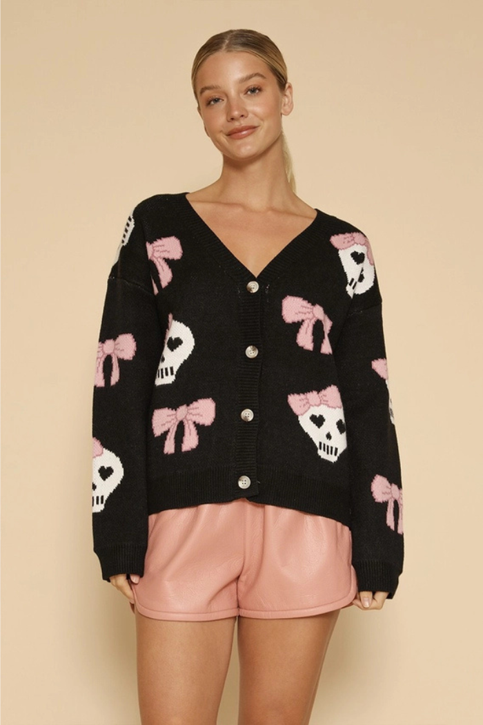 A woman wears a black v-neck cardigan with white skull and pink bow intarsia patterns, paired with light pink shorts.