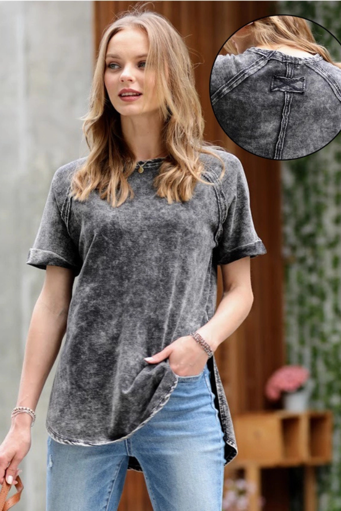 A woman wearing a black acid-wash short-sleeved top with a round neck, cuffed sleeves, and a high-low hem with side slits, paired with light-wash jeans. A close-up inset shows the detail of the double back seam and textured fabric on the back of the top.
