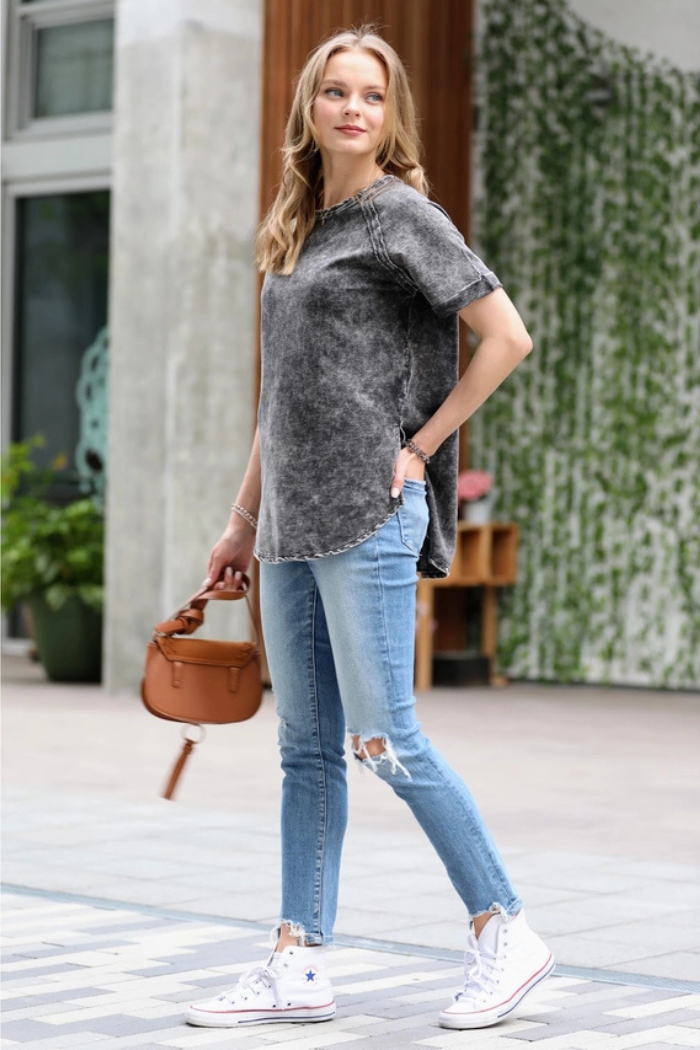 A woman with long, blonde hair wearing a black acid wash t-shirt, light-wash ripped skinny jeans, and white high-top Converse sneakers. She is carrying a small, brown leather handbag and is walking on a paved surface outdoors with a building in the background.