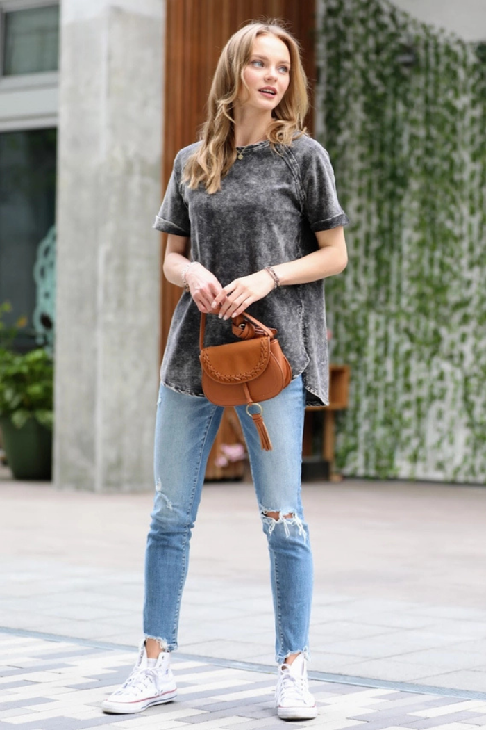 A woman with blonde hair wearing a black acid wash t-shirt, ripped light-wash jeans, and white high-top sneakers, holding a small brown saddlebag purse. She is standing outdoors with a slight smile, looking towards the right side of the frame.