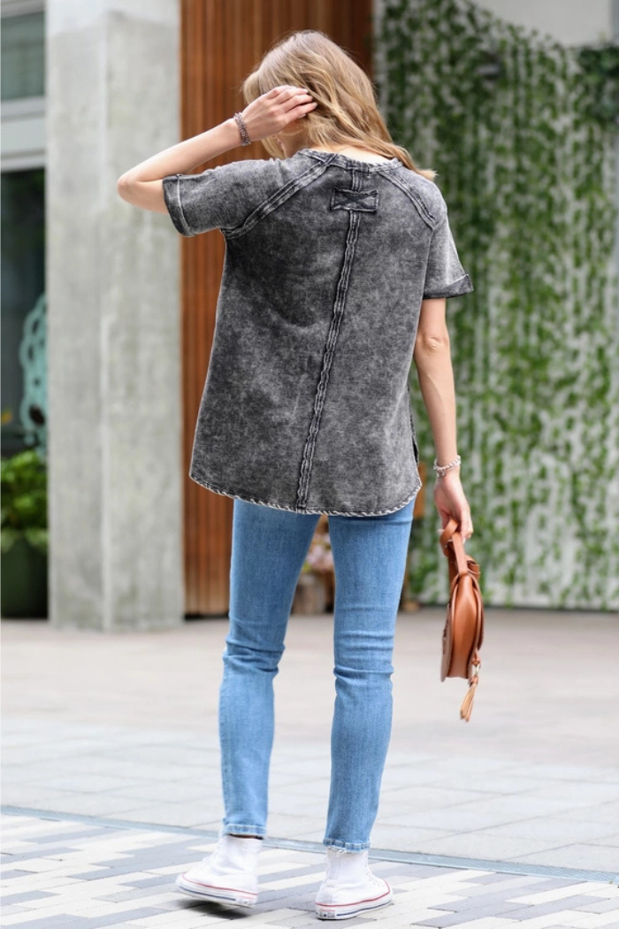 A woman from behind, wearing a black acid wash t-shirt, light blue skinny jeans, and white sneakers with white socks. She is holding a brown handbag in her left hand and her right hand is near her head. The background shows a building with vertical wooden slats.
