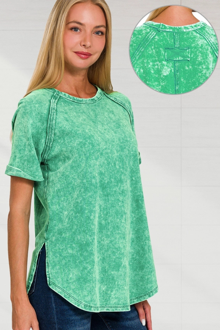 A woman wearing a green acid-wash, short-sleeved top with visible seams and a rounded hem, paired with dark jeans. A close-up inset shows the detailed stitching and back patch of the top near the neckline.