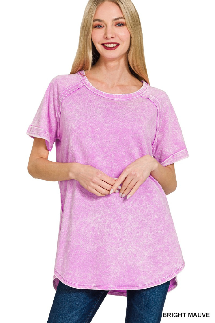 A woman models a short-sleeved, round neck pullover in a bright, acid-wash pink or mauve color. The shirt has a relaxed fit and a rounded hemline, with visible seam details on the shoulders and back. The fabric appears to be French terry, giving it a textured look.