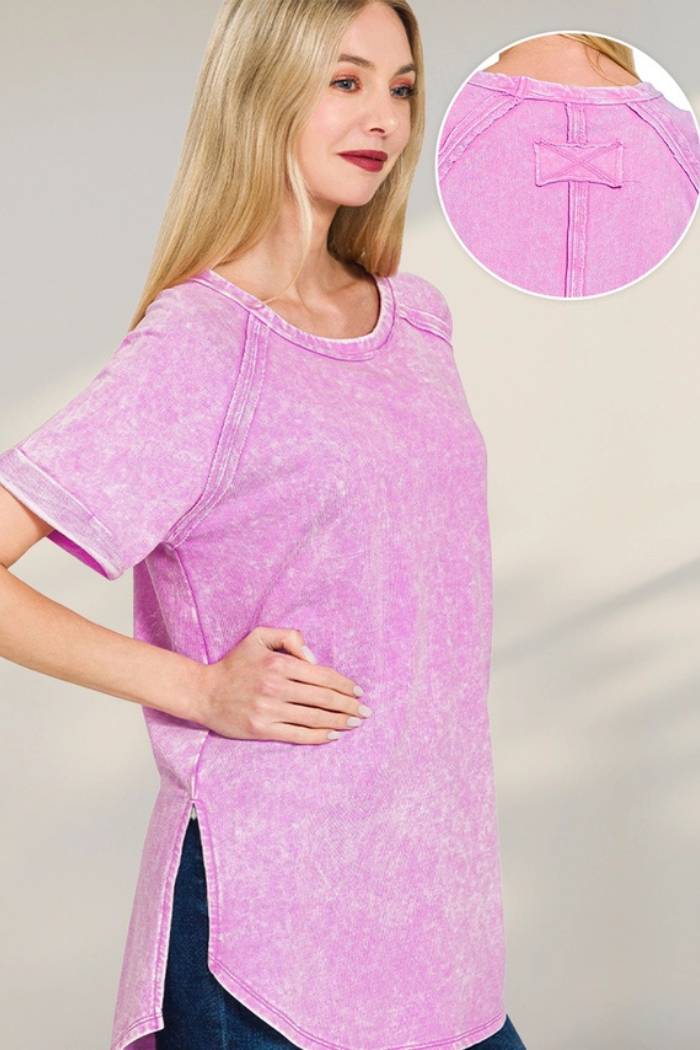 A woman wearing a pink acid wash short-sleeved top with a rounded hem and side slits stands with her hand on her hip, looking towards the left. An inset image in the upper right shows a close-up of the back neckline of the top, revealing a small decorative fabric patch.