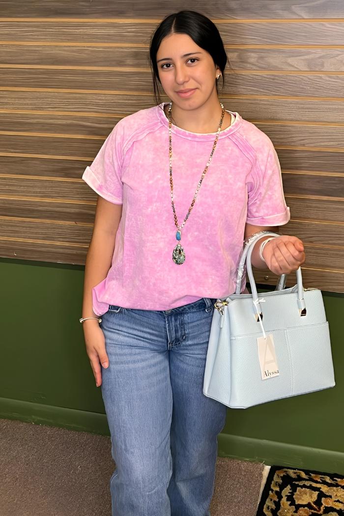 A young woman with dark hair stands in front of a horizontally paneled wooden wall, wearing a light pink acid wash, short-sleeved t-shirt, blue jeans, and a long, beaded necklace with a prominent pendant. She is holding a light blue handbag with a tag that reads "Alyssa".