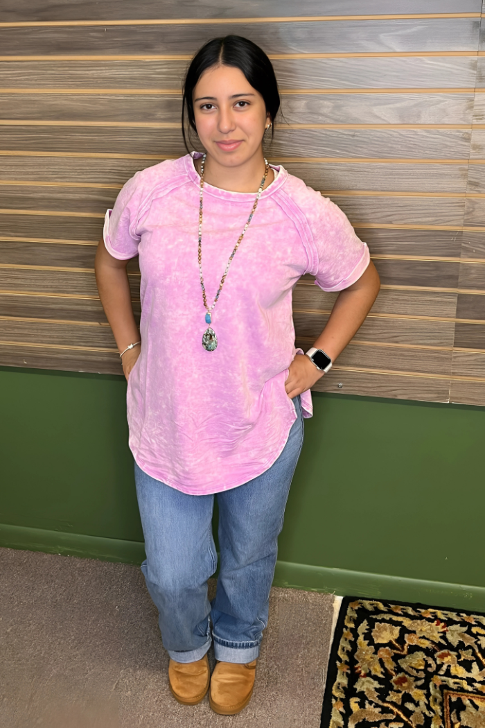 A young woman with dark hair stands facing forward, wearing an acid wash pink t-shirt, light-wash jeans, and tan shoes. She has a long-beaded necklace with a prominent pendant and a smartwatch on her left wrist. She is smiling slightly, and her hands are in her pockets. The background appears to be a wooden wall, and a patterned rug is visible at the bottom right.