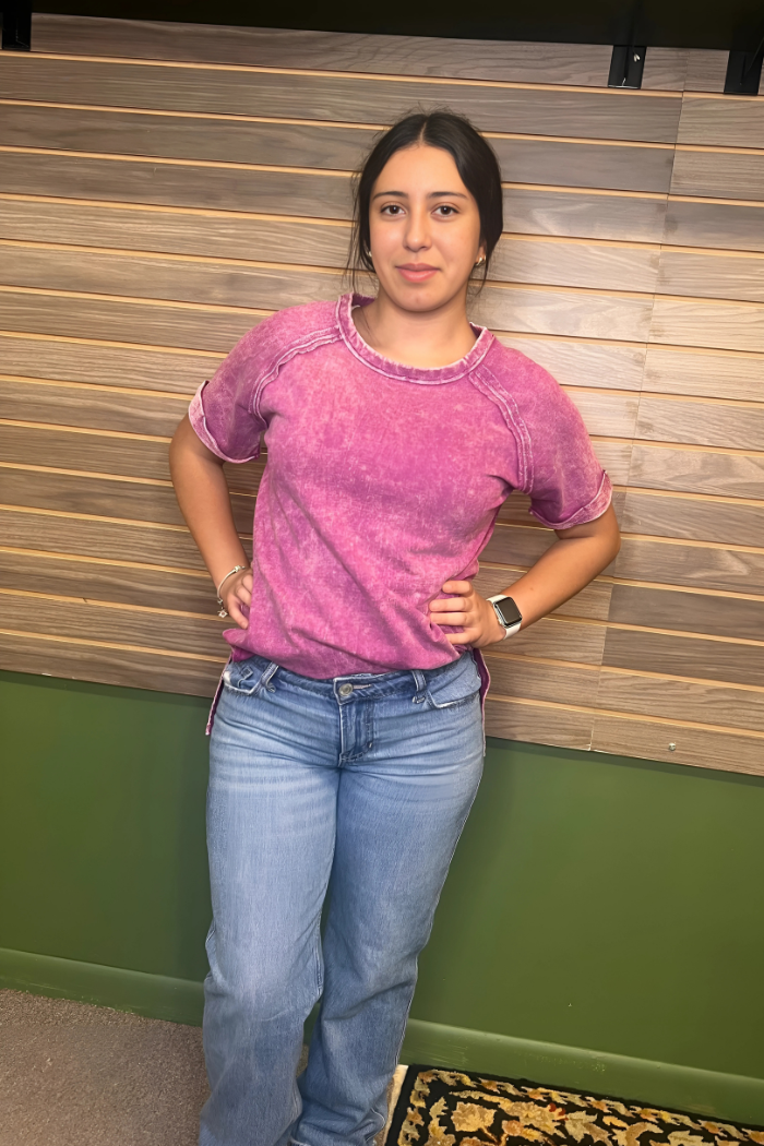 A young woman with dark hair wearing an acid wash plum short-sleeved top and light blue jeans. She has her hands on her hips and is looking directly at the camera.