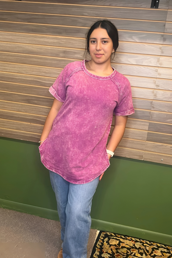A young woman with dark hair wearing a short-sleeved, acid-wash plum French terry top with a round neck, cuffed sleeves, and a rounded hem, paired with light-wash blue jeans.