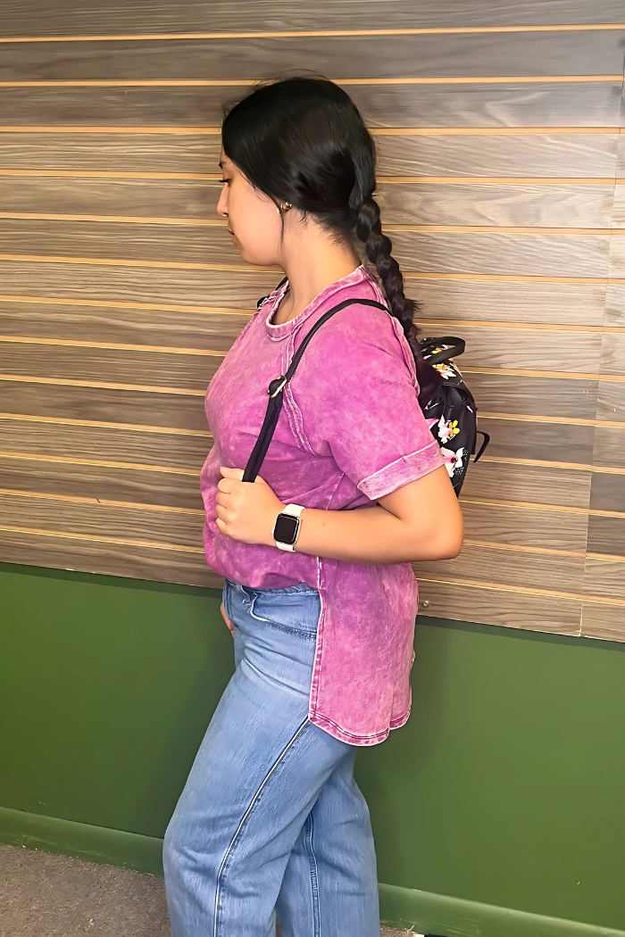 A young woman with dark hair styled in a braid, wearing an acid wash plum t-shirt, light blue jeans, a white smartwatch on her left wrist, and a black floral backpack slung over her right shoulder.