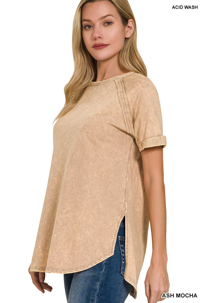 A woman wearing a tan/beige color acid wash French terry short-sleeve top with raglan sleeves, cuffed sleeves, and a rounded, raw-edge hem with side slits. She is also wearing blue jeans. The top has visible double back seams.