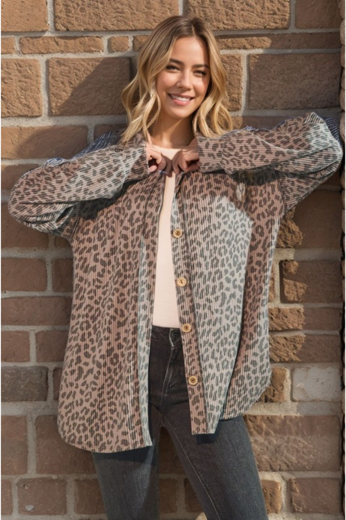 A woman with blonde hair smiles at the camera, wearing a button-up shirt with taupe and black animal print, over a white top and dark jeans, standing against a brick wall.