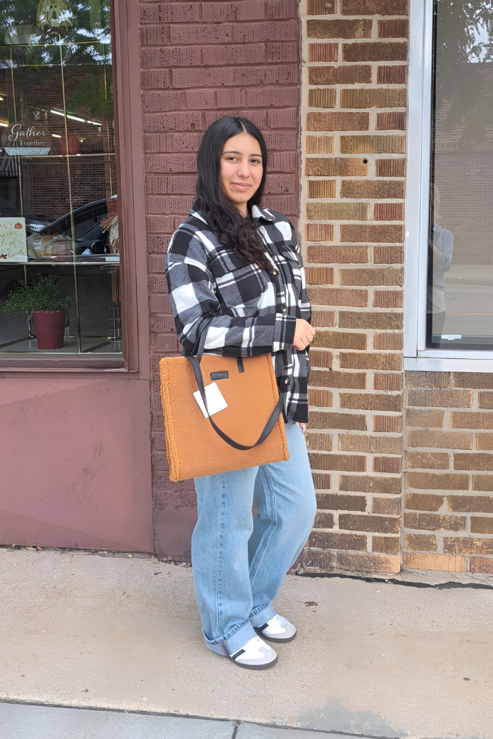 A woman with dark hair stands outdoors in front of a brick building and a storefront window, smiling slightly. She is wearing a black and white checkered shacket, light blue jeans, and light-colored shoes. She is holding a large, brown, textured tote bag with a black handle in her left hand.