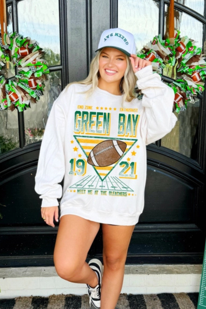 A blonde woman smiling and wearing a white baseball cap with "GREEN BAY" written in green, and a white long-sleeved sweatshirt. The sweatshirt features a distressed vintage-style graphic with "END ZONE ENTHUSIAST" at the top, "GREEN BAY" in large green letters, a football over a stylized field with the numbers "19" and "21" on either side, and "MEET ME AT THE BLEACHERS" at the bottom.
