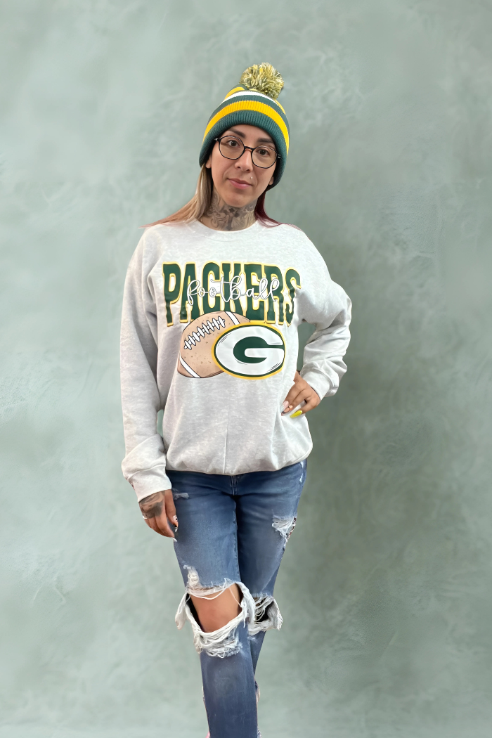 A woman is posing while wearing a light gray Green Bay Packers crewneck sweatshirt, ripped blue jeans, glasses, and a green, yellow, and blue striped beanie with a pom-pom. The sweatshirt features "PACKERS" in green lettering above a football and the Packers "G" logo.