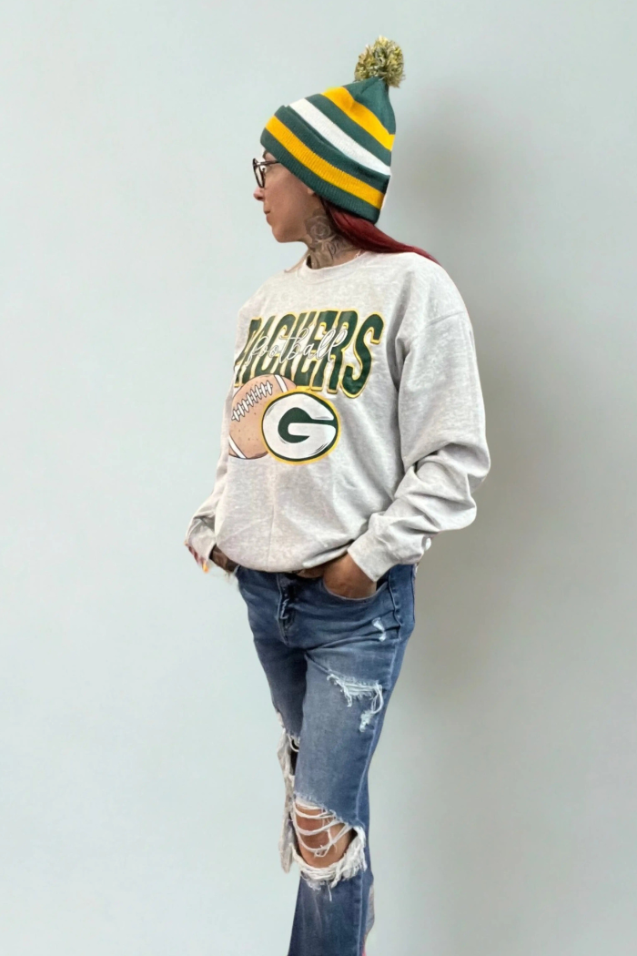 A woman, seen from the side, wearing a cream-colored Green Bay Packers sweatshirt featuring the team's logo with a football graphic and "PACKERS" text, distressed blue jeans with large rips at the knees, and a green, yellow, and white striped beanie with a pom-pom. The person also wears glasses and has hands in their pockets.