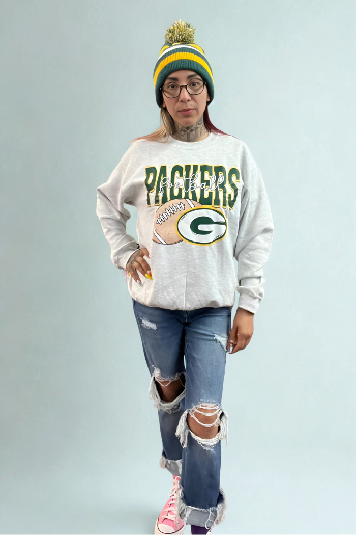 A woman stands posing, wearing a light grey Green Bay Packers oversized crewneck sweatshirt with a football graphic and the team's "G" logo, distressed blue jeans with large rips at the knees, and pink high-top Converse sneakers. She is also wearing a green, yellow, and white Green Bay Packers beanie with a pom-pom on top and clear-framed glasses. Her right hand is placed on her hip. The background is a plain light blue wall.