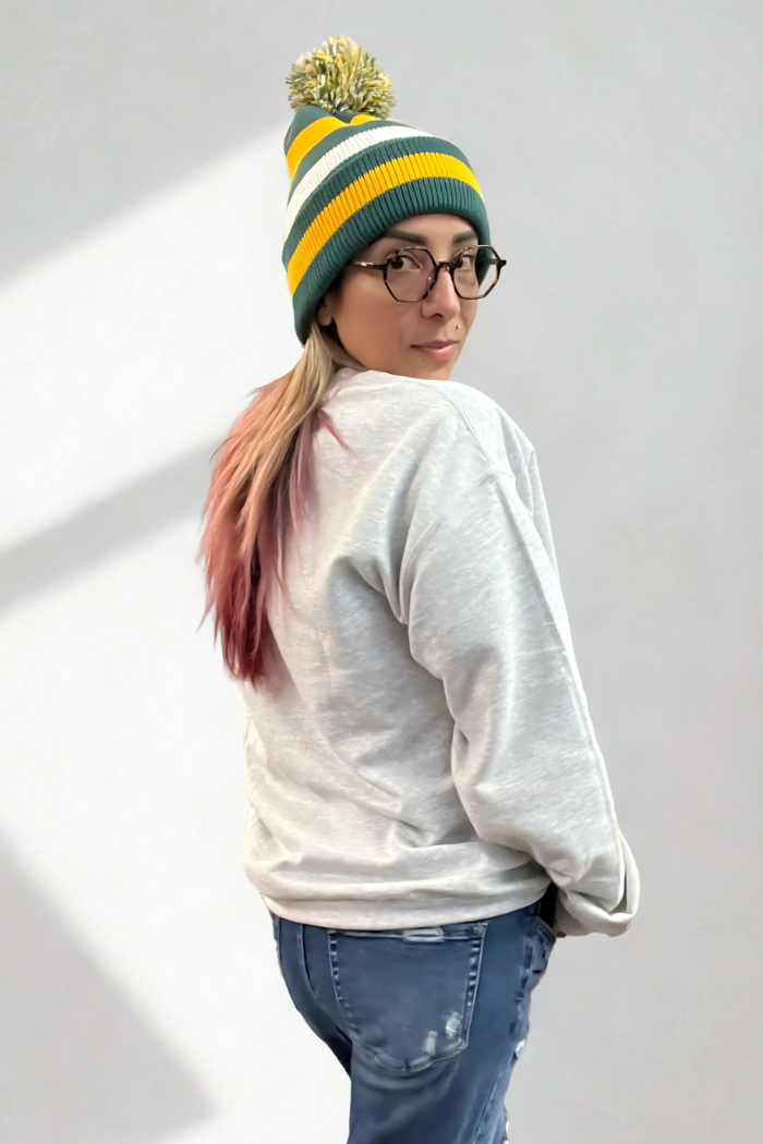 A woman with pink-tipped hair in a ponytail wears a green and yellow striped beanie with a pom-pom, round glasses, with back view of the grey sweatshirt, and blue jeans, looking back over her shoulder with a slight smile against a white background.