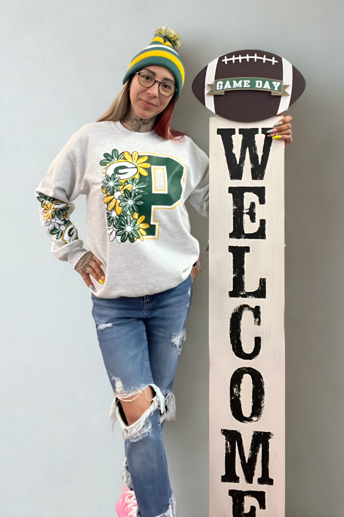 A woman wearing a Green Bay Packers-themed sweatshirt with floral accents, ripped jeans, and a matching beanie poses next to a tall, white wooden "WELCOME" sign topped with a football that says, "GAME DAY".