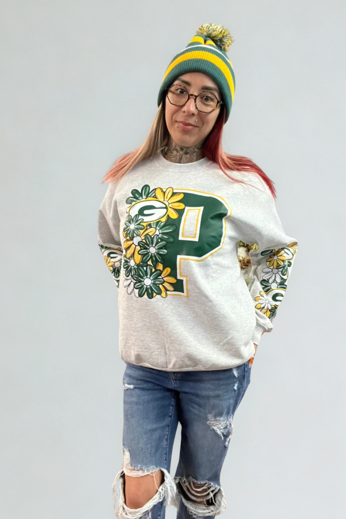 A person wearing a grey Green Bay Packers sweatshirt with a large floral "P" logo, distressed blue jeans, and a green and yellow striped knit beanie with a pom-pom, stands against a plain grey background. The sweatshirt sleeves also feature floral patterns.