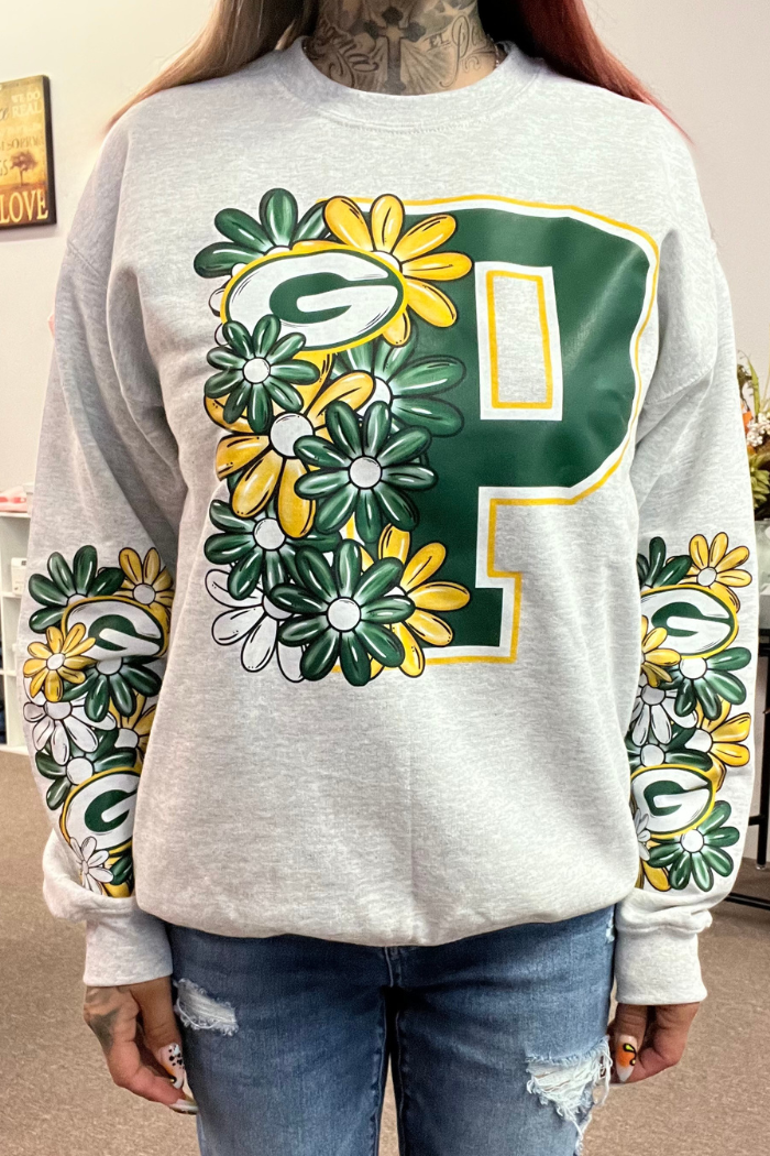 A person wears a light grey sweatshirt featuring a large Green Bay Packers logo (a green 'P' with a smaller 'G' inside) adorned with green, yellow, and white floral designs. Similar floral patterns with the Packers logo are also present on the sleeves."