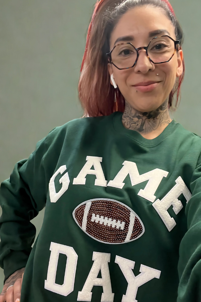 A woman with red-streaked hair, glasses, and visible neck tattoos smiles at the camera, wearing a green sweatshirt with "GAME DAY" written in white chenille letters and a brown football patch in the center.