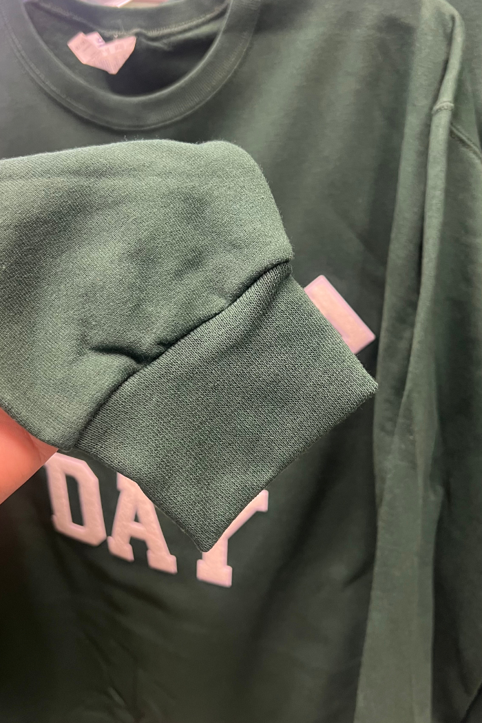 A close-up shot of a dark green long-sleeved sweatshirt, showing the ribbed cuff of the sleeve and part of the main body. White lettering, partially visible, spells out "DA" and what appears to be part of another word or letter.