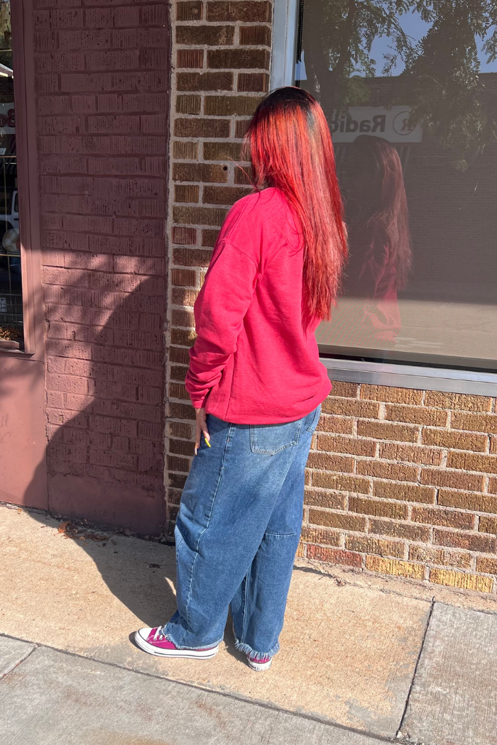 A person with long, bright red hair, wearing a red sweatshirt, baggy blue jeans, and pink low-top sneakers, stands on a sidewalk next to a brick building. The person is viewed from the side, looking away from the camera. The reflection of a sign is visible in the building's window.