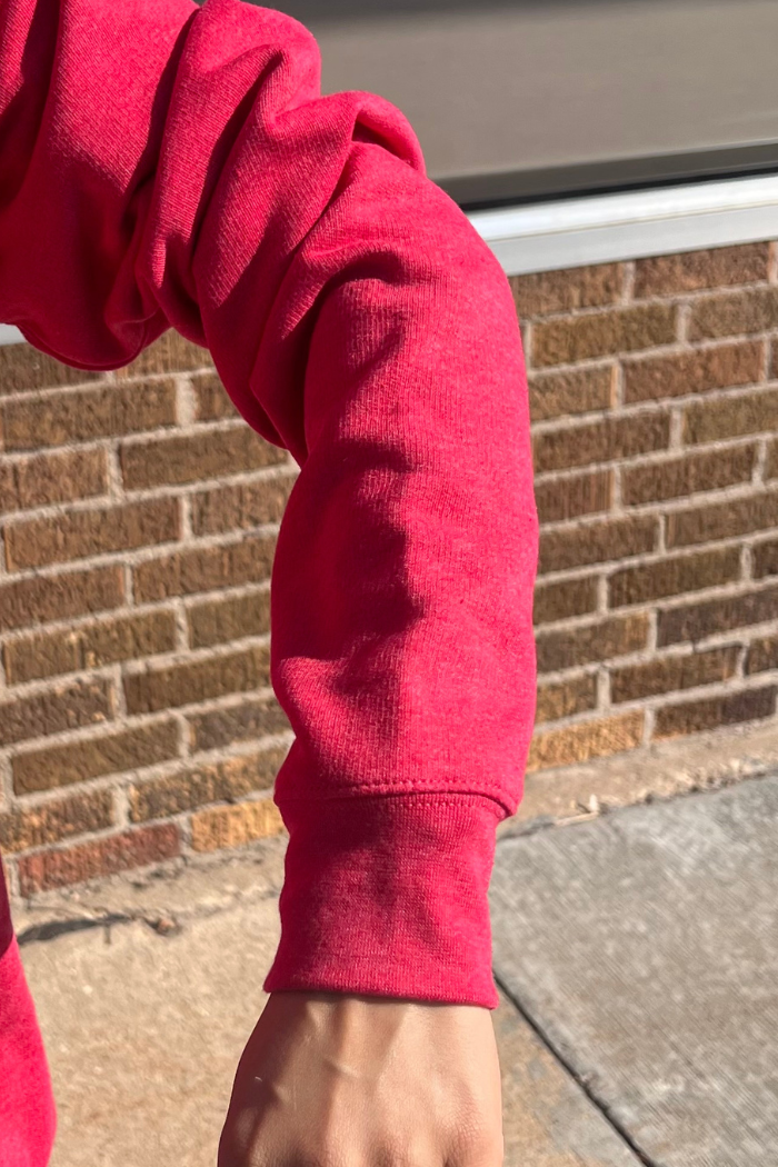 A close-up shot of a person's bent arm wearing a red sweatshirt with a ribbed cuff at the wrist. The background is a brick wall.