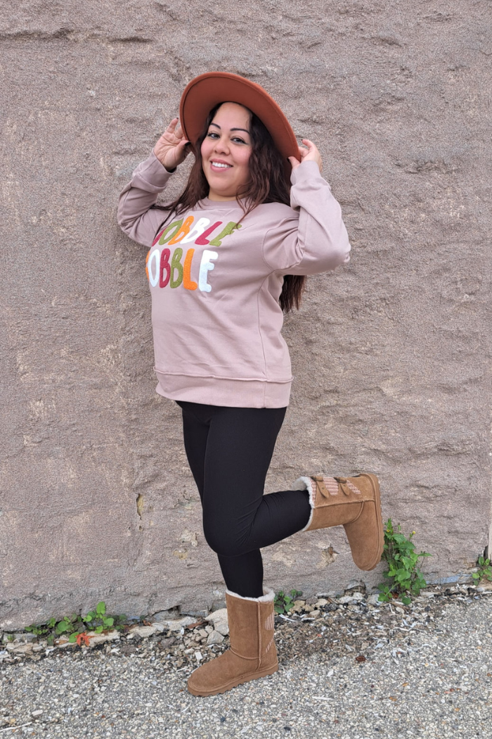 A young woman with dark hair and a wide smile poses in a beige sweatshirt with the word "GOBBLE" on the front in colorful letters, black leggings, and tan boots with fur trim. She is holding the brim of her rust hat with both hands while lifting one leg. The background is a textured grey stone wall and a concrete ground with some green vines growing on it.