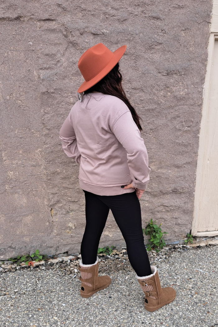 A woman wearing a dark khaki oversized sweatshirt, black leggings, and brown boots stands with her back to the camera. She is wearing a wide-brimmed hat. The background is a textured gray stone wall.