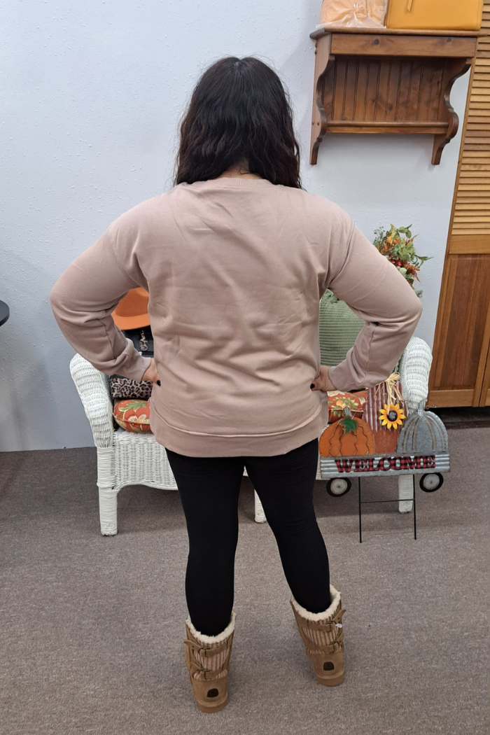The image shows a woman from behind, with long dark hair, wearing a dark khaki colored sweatshirt, black leggings, and tan-colored Ugg-style boots. She is standing with their hands on their hips in what appears to be a retail store or boutique, with a white wicker chair and a wooden shelf visible in the background. There is also a "WELCOME" sign on a metal stand shaped like a pumpkin and decorated with sunflowers.