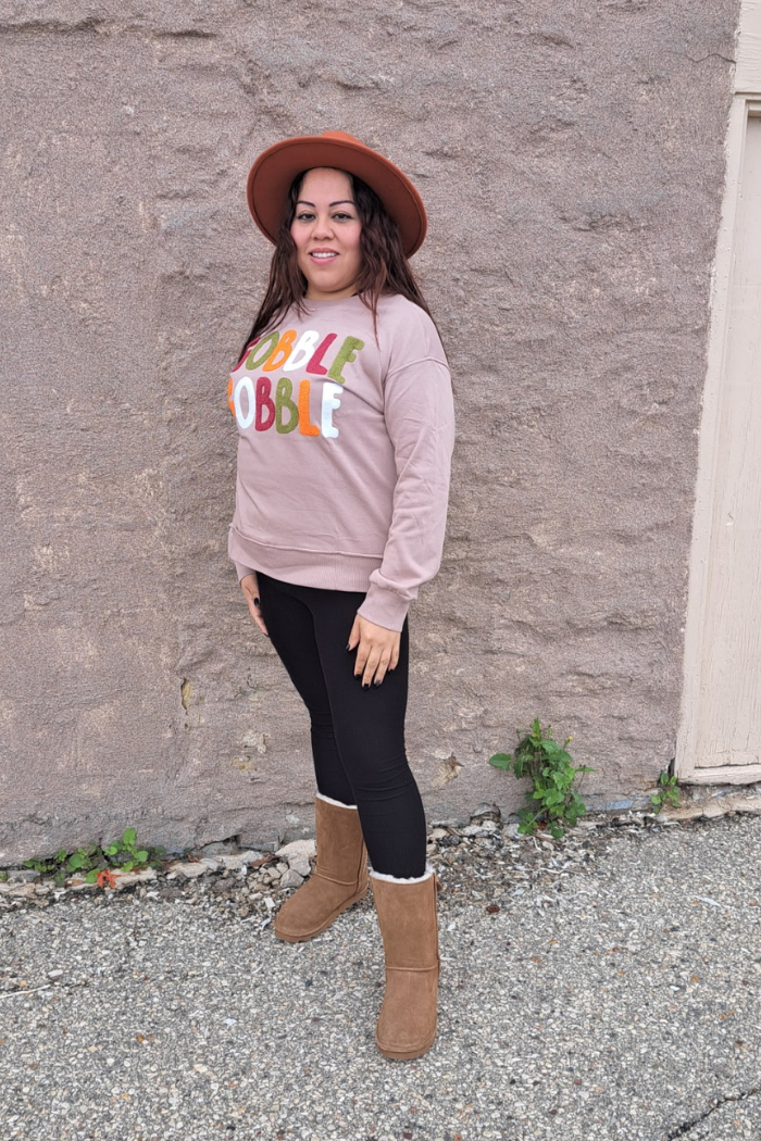 A woman wearing a Thanksgiving-themed sweatshirt, a rust hat, black leggings, and brown boots. She is standing outdoors against a stone wall. The sweatshirt has the text "GOBBLE GOBBLE" on it in colorful letters.