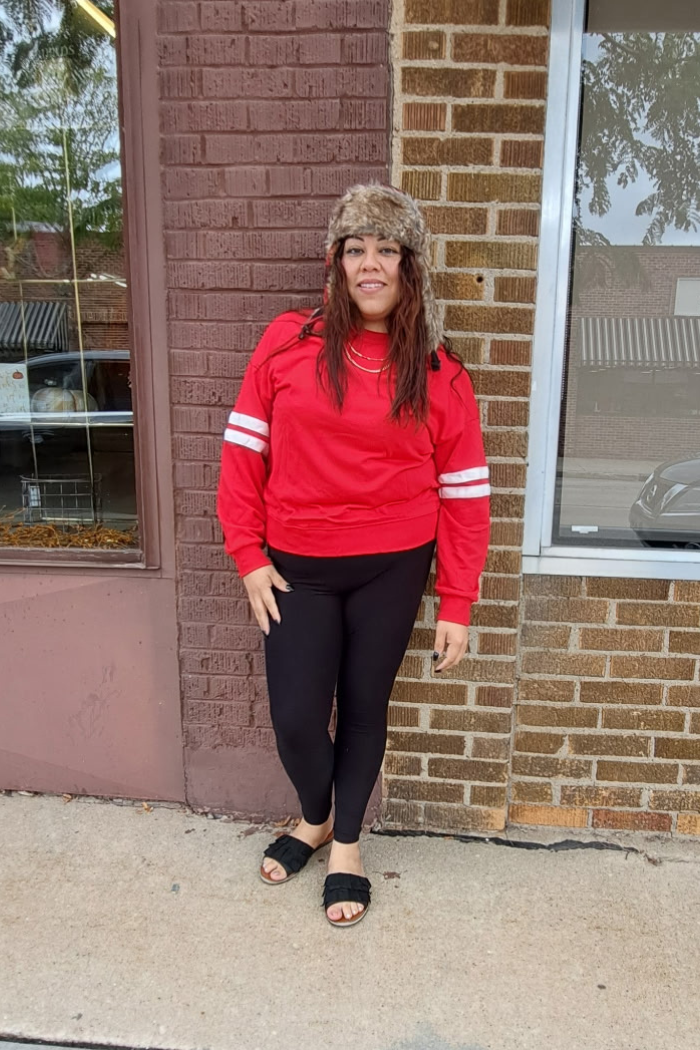 A woman wearing a red sweatshirt with white stripes on the sleeves, black leggings, black slide sandals, and a furry brown hat stands on a sidewalk in front of a brick building.