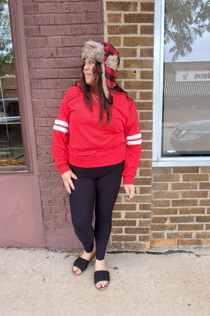 A woman stands in front of a brick wall, wearing a red sweatshirt with white stripes on the sleeves, black leggings, and black fuzzy sandals. She is also wearing a red and black plaid trapper hat with a fur trim.