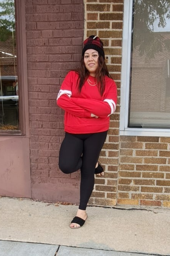 A woman with brown hair, wearing a red long-sleeved sweatshirt, black leggings, and black slides, stands leaning against a brick wall. She is also wearing a patterned beanie and a necklace, and has her arms crossed.