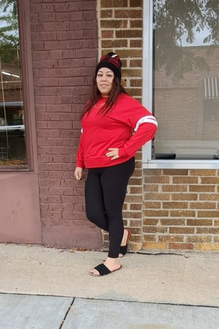 A woman with brown hair stands outdoors, leaning against a brick wall. She is wearing a red oversized sweatshirt with white stripes on the sleeves, black leggings, and black sandals.