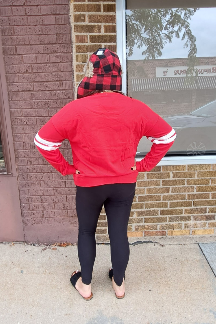 A person from behind wears a red sweatshirt with white stripes on the sleeves, black leggings, and a red and black buffalo plaid hat.