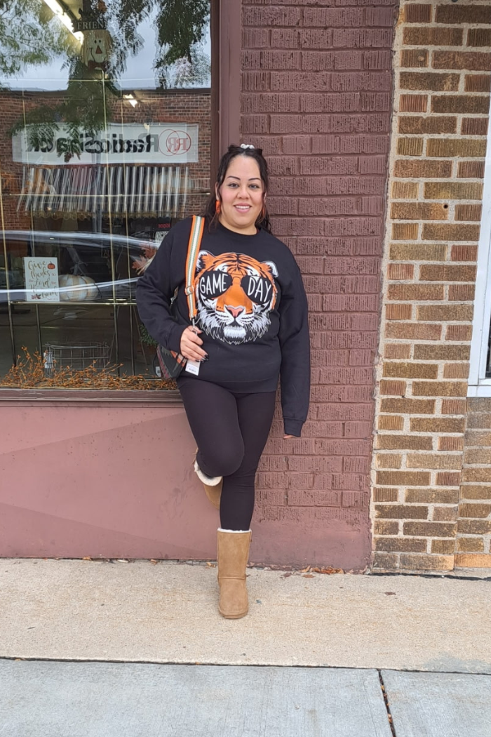 A woman stands smiling and leaning against a brick wall and a window. She is wearing a black sweatshirt with a graphic of a tiger wearing sunglasses that say, "GAME DAY". She is also wearing black leggings and brown boots and has a bag with an orange and white striped strap.