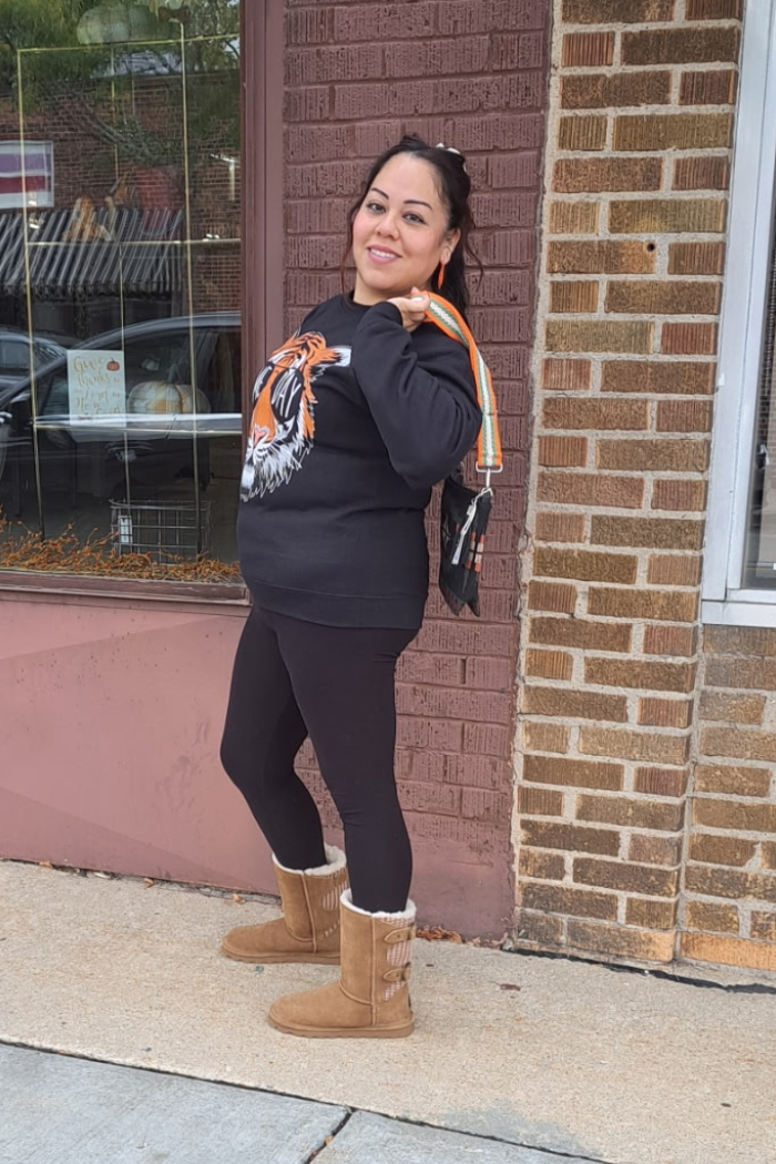 A woman with dark hair in a half-up ponytail is standing outside a brick building with a large window. She is wearing a black sweatshirt with a tiger head design, black leggings, and brown boots. She is also carrying a shoulder bag with a patterned strap.