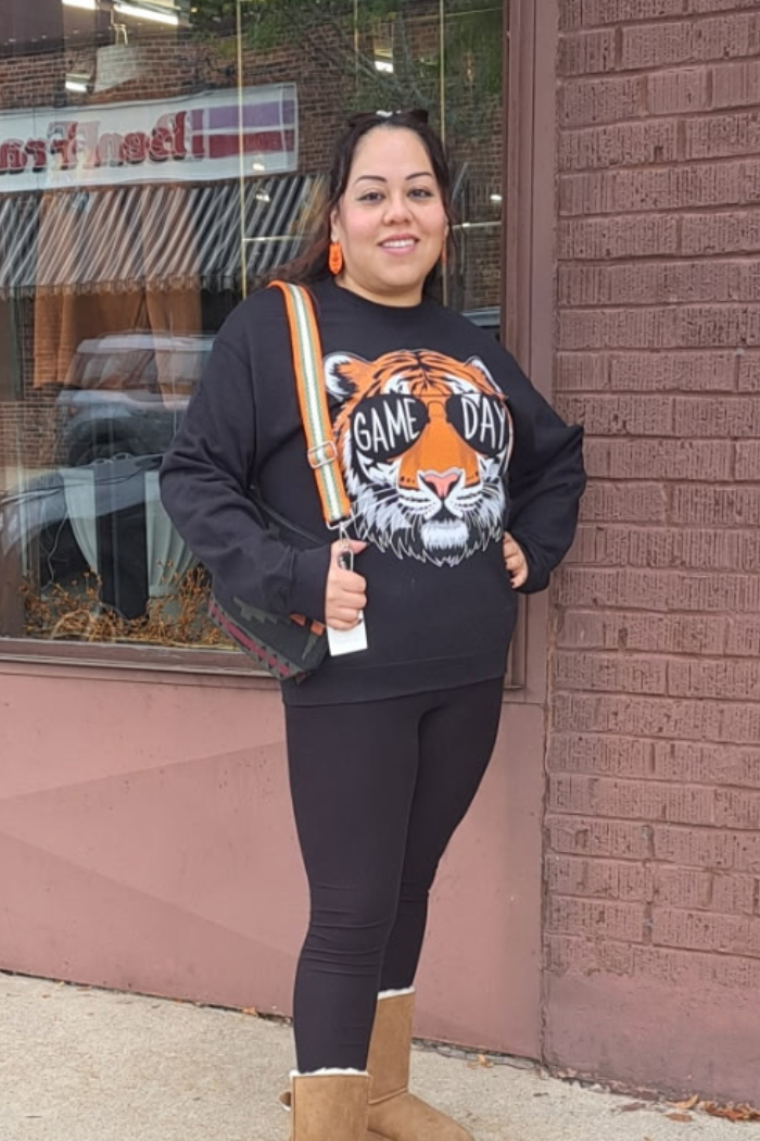 A woman standing outdoors wearing a black sweatshirt with a graphic of a tiger's head wearing sunglasses and the text "GAME DAY" on the front. She is also wearing black leggings and a striped bag over her shoulder.