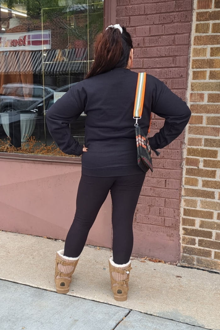 A person stands with their back to the camera, wearing a black long-sleeved sweatshirt and black leggings. She has her hands on their hips. The person is wearing tan, fur-lined boots and a colorful strap bag with an orange, green, and white stripe pattern. The setting appears to be a brick building with a large window, reflecting the surroundings.