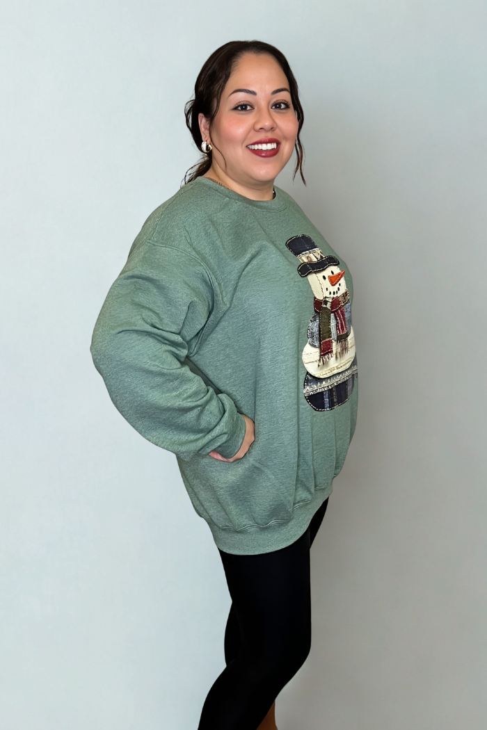 A woman smiles as she poses sideways in the Snowman Patchwork Holiday Sweatshirt, a cozy sage green top with a snowman graphic, paired with black leggings and set against a plain light gray background.