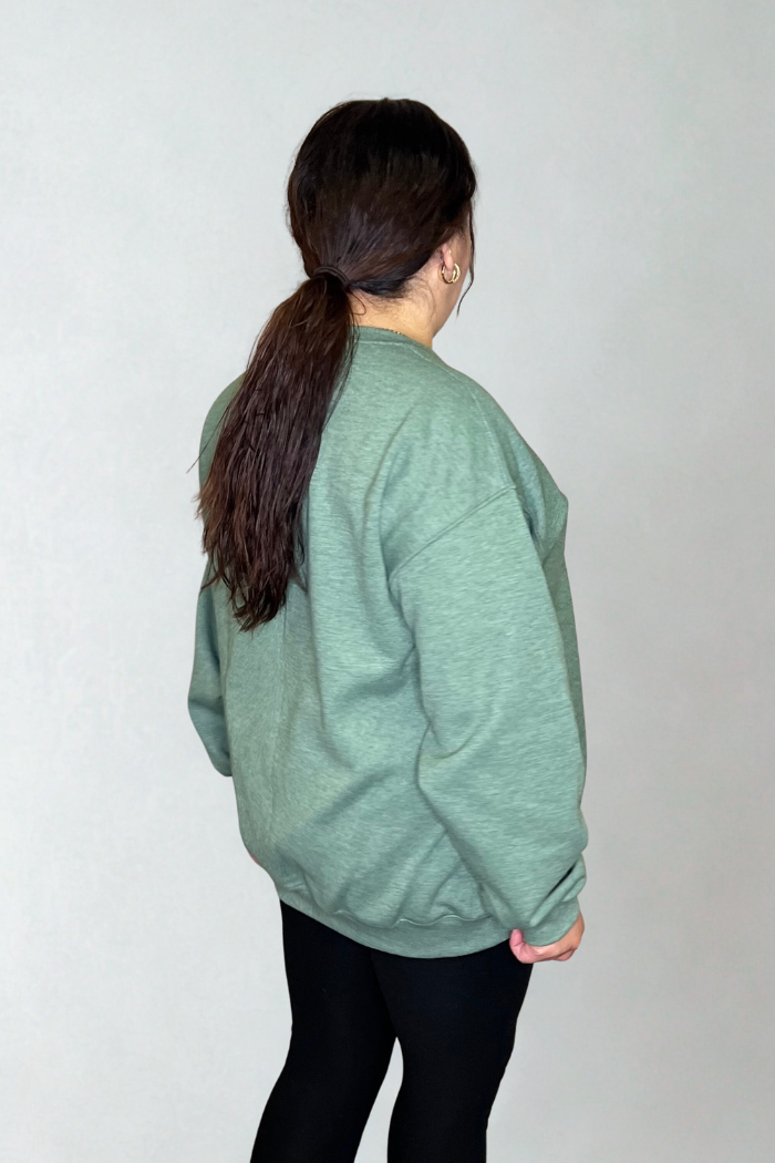 A person with long brown hair in a ponytail, wearing the Snowman Patchwork Holiday Sweatshirt and black leggings, stands facing away from the camera against a plain light background.