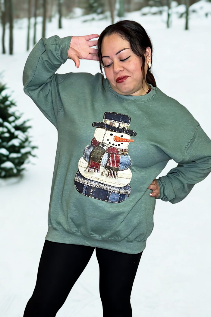 A woman in a Snowman Patchwork Holiday Sweatshirt and black leggings poses playfully outdoors, surrounded by snow and trees.