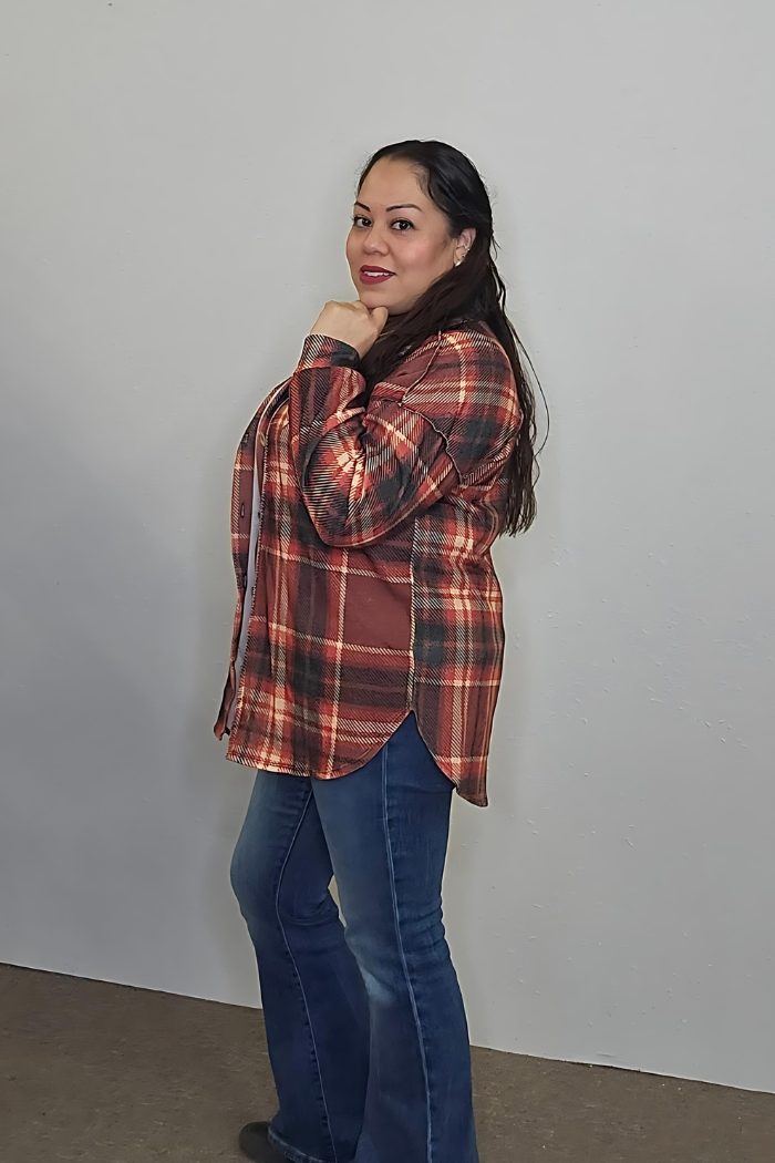 A woman with long dark hair, wearing the Plaid Flannel Shacket and blue jeans, stands sideways against a light wall, smiling softly and resting her chin on her hand—an ideal choice for easy fall layering.