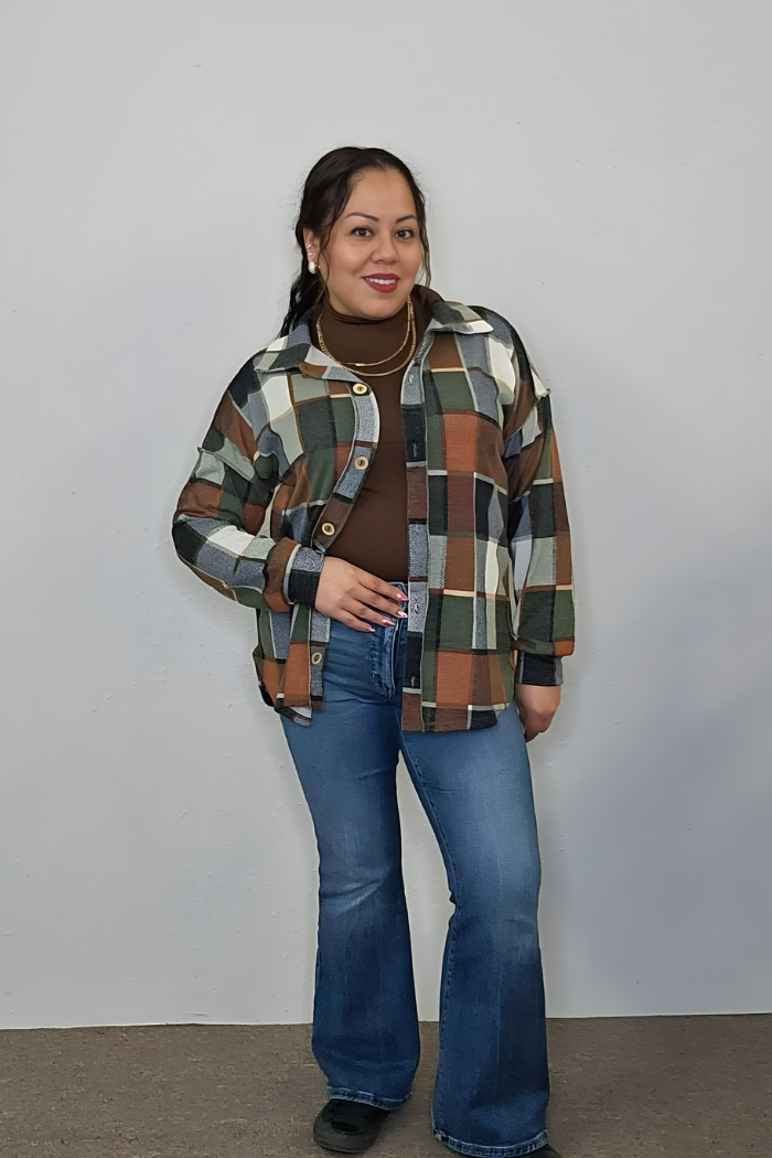 A woman stands against a light wall, wearing a brown turtleneck, the Plaid Print Shirt Jacket in earth tones, blue flared jeans, and black shoes, with one hand on her hip and a subtle smile.